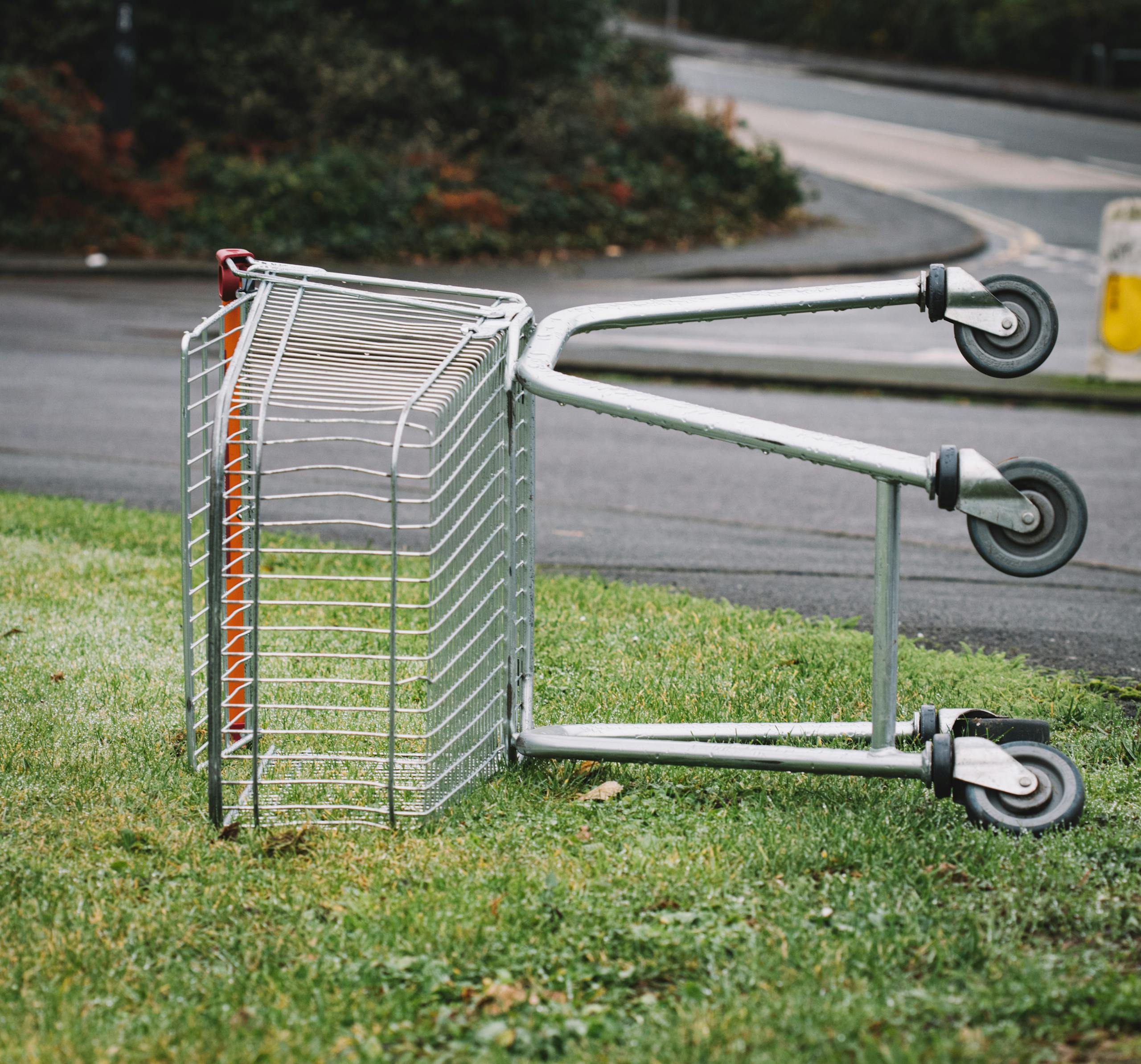 An empty shopping cart lies abandoned on a patch of grass by the roadside.