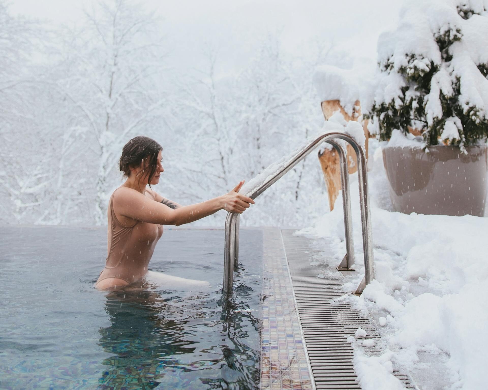 A woman in a swimsuit enters a heated pool surrounded by snow-covered landscape in winter.
