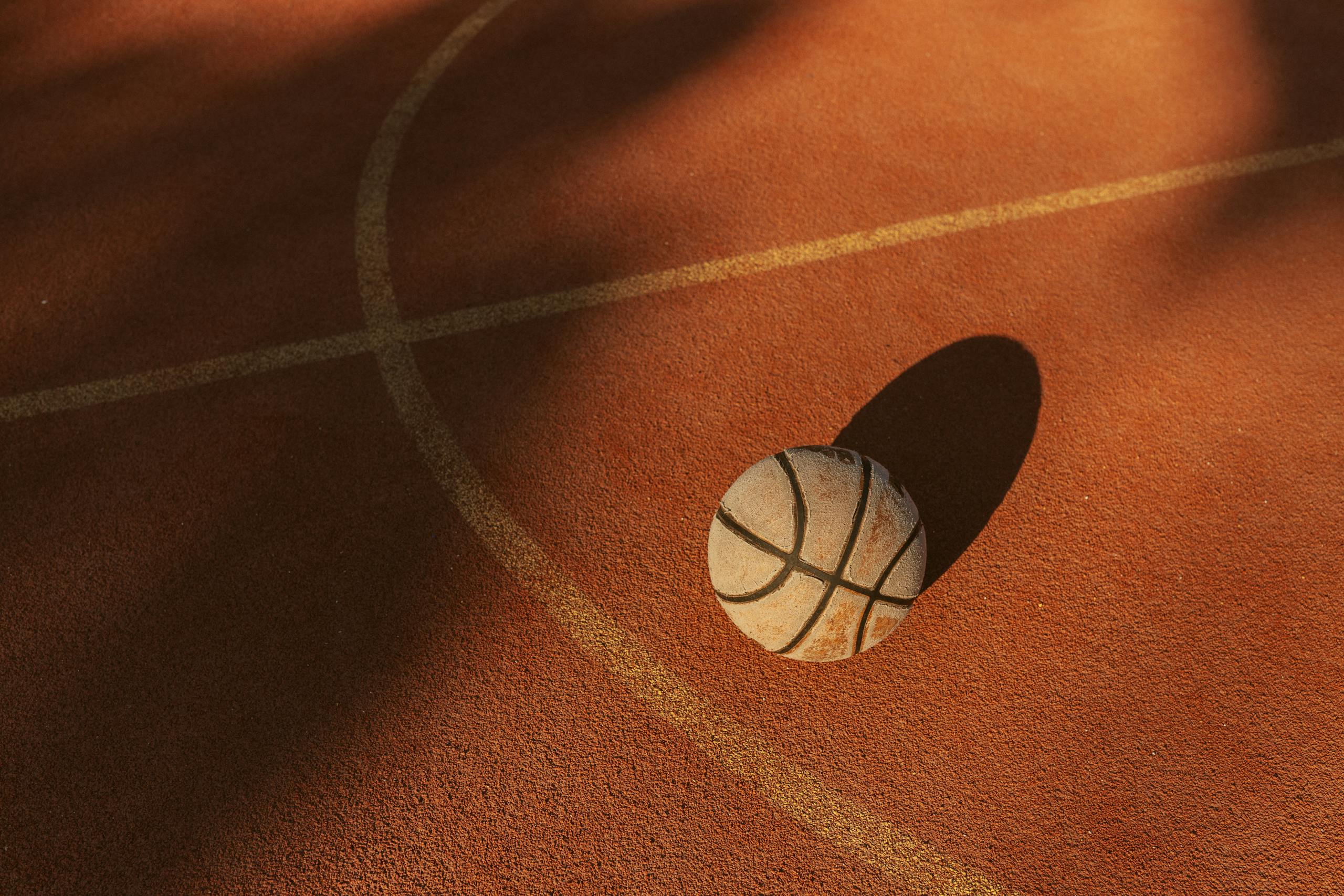 A basketball sits alone under sunlight creating bold shadows on an outdoor court.