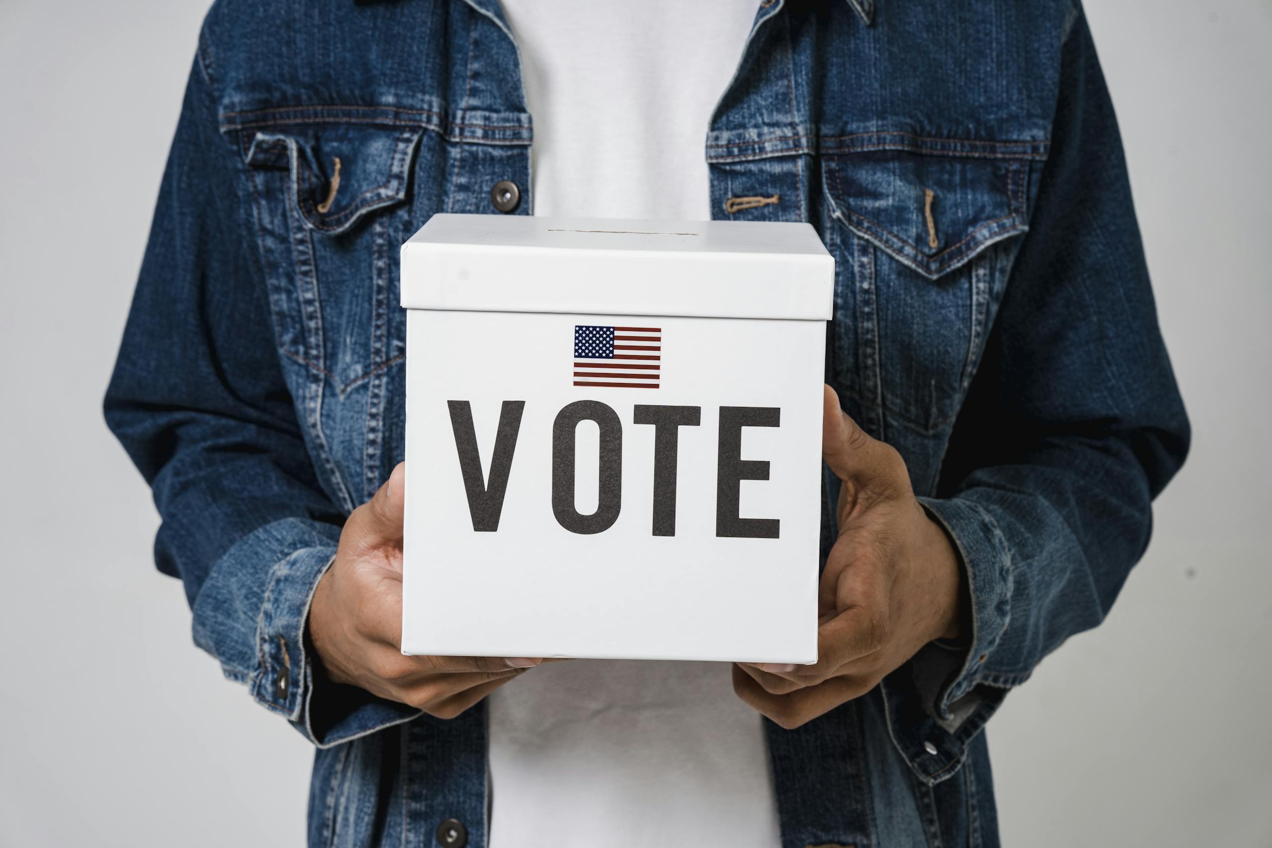 Person in denim holding a ballot box with a US flag, emphasizing voting rights.
